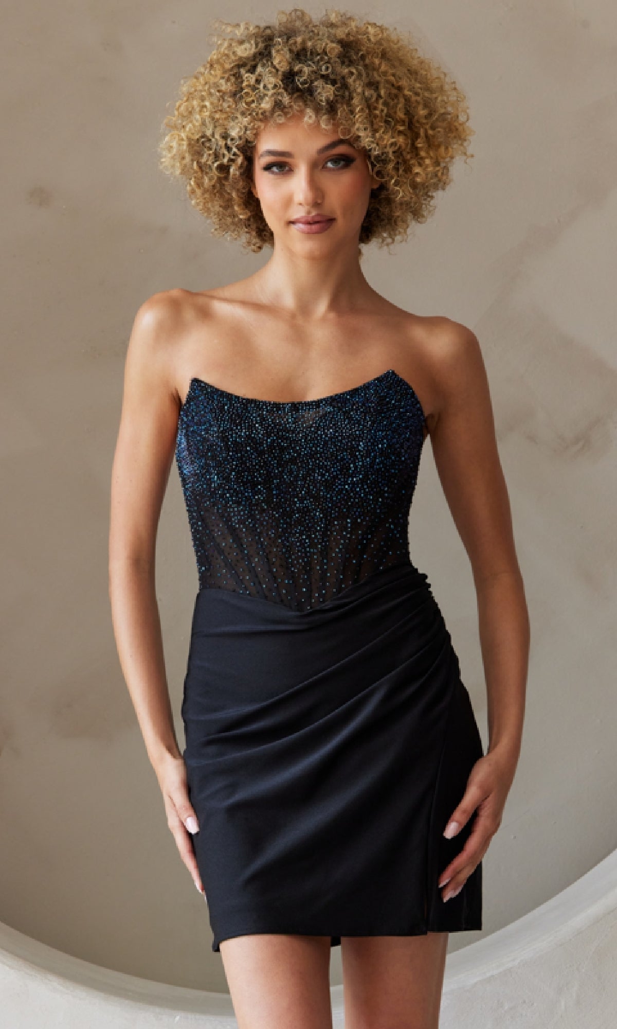 A woman in the Beaded-Bodice Strapless Short Hoco Dress 3021S, featuring a sparkly beaded corset bodice and a ruched black mini skirt, stands against a neutral background.